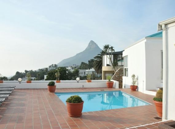A sunny pool terrace beside a white house, with mountains behind